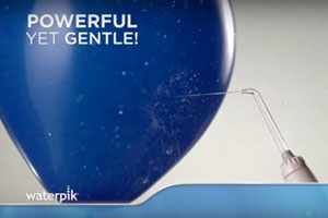 Blue balloon with the words Powerful Yet Gentle! with a Waterpik® Water Flosser handle and tip shooting water against it