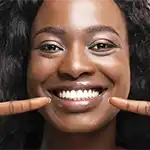 Woman with dark curly shoulder length hair smiling and pointing to her teeth