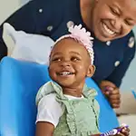 Young child with pink hairband and green dress, smiling, holding a toothbrush, with woman behind her smiling
