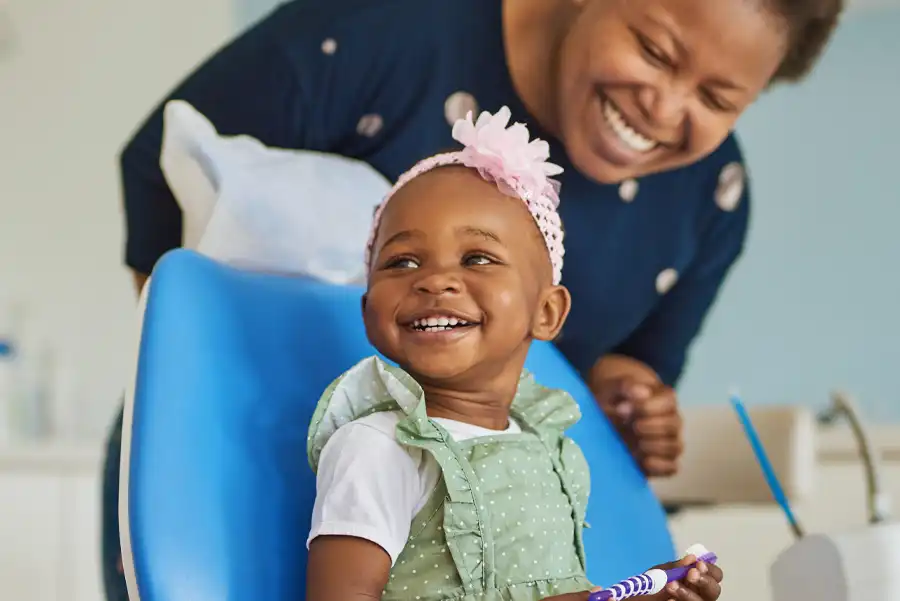 Young child with pink hairband and green dress, smiling, holding a toothbrush, with woman behind her smiling