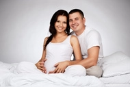 Expectant parents sitting together on a bed, with the pregnant woman holding her belly and her partner embracing her, both smiling.