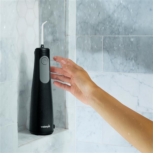 Black Cordless Enhance 3200 water flosser on a shower ledge, with a hand reaching for it.