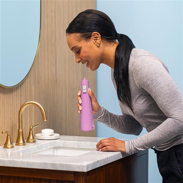 Mauve Cordless Pulse 3100 handheld water flosser in use, with person leaning over the bathroom sink.